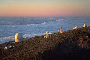 Series of white structures along the side of a mountain with a sea of clouds below and behind the mountain extending to the horizon which is red, orange, and yellow.