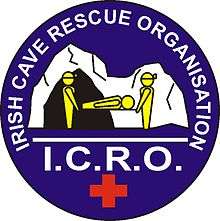 Circular graphic logo, depicting two cavers carrying a casualty on a stretcher away from a cave entrance.