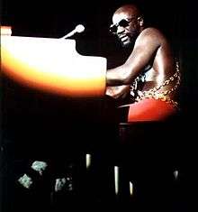 An adult male with sunglasses plays a piano under a spotlight on a darkened stage, 1973