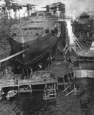 The hull of a large ships in a shipyard, surrounded by scaffolding and cranes