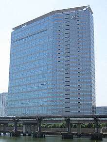 A modern multi-storey building in blue and grey colour, with Japan Airlines' "JAL" logo on the top right, there are blue sky on the background and a highway bridge in the foreground