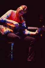 A color photograph of Jaco Pastorius sitting on a stool and playing a bass guitar