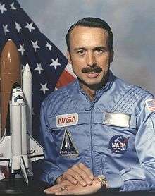 man with mustache in astronaut uniform, model of space shuttle to side, U.S. flag in background
