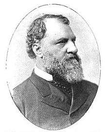 Profile of a white man with curly hair, a receding hairline, and a full beard, wearing a suit coat over a shirt and tie.