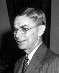 Head and shoulders of smiling man in suit and tie with round dark-rimmed glasses. This is a detail from the picture below.