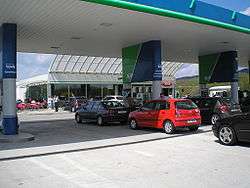 Cars queuing up at a filling station. A typical motorway rest area café and convenience store are visible in the background.