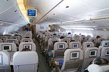 Inside view of an aircraft's Economy Class cabin with television on the back of the seats and overhead lockers on the ceiling