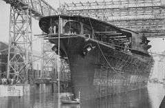 A large ship, covered with scaffolding, sits in harbor under a large gantry.