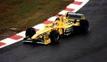 A figure, wearing a helmet with a white, red, yellow and blue design, is driving a Formula One that is of a yellow and black colour scheme.