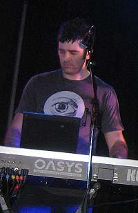 Upper body shot of a 34-year-old man. He is partly obscured by a keyboard and microphone in front of him. He has short dark hair and wears a dark tee-shirt which has a large open eye logo. His eyes are directed down and slightly to his right. His arms are raised to the keyboard but his hands are obscured. The lettering O-A-S-Y-S is visible on the back of the synthesiser with an additional K at the right.