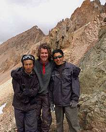 Three people stand outside facing the camera, mountaintops are in the backdrop, the tallest bearded Western man in the middle has his arms around the shoulders of two shorter Asian men.