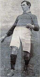 A black and white photograph of a man wearing a dark coloured football shirt and white shorts.