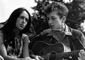 A black and white photograph of Joan Baez and Bob Dylan singing while Dylan plays guitar