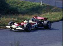 Side-view color photograph of Rindt racing a red and white Lotus Formula One car with a wing attached at the rear