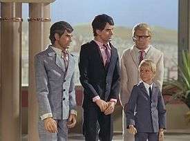 Three men and one boy stand in a palatial setting. A desert landscape is visible from a balcony in the background. One man on the far left is grey-haired and wears a grey suit and tie, the man to the right of him dark-haired and in deep navy blue. Both men are orientated in the direction of the (blond-haired) boy, who is also formally attired in grey. The third man, also blond but wearing a cream-shaded suit, stands directly behind him.