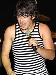 A male with short black hair. The male is wearing a black and white tank top and he is holding a microphone.