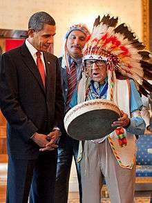 An old man in full feathered headdress plays a drum with a man in a suit watching