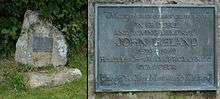 A headstone made of an irregularly-shaped rock, with a lead plaque on it, in front of a hedge