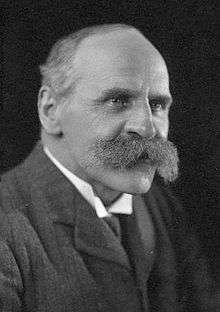  A three-quarter view from the left monochrome portrait of a middle-aged white man with a receding hairline and a bushy moustache. He is wearing a jacket and a stiff white collar.