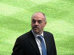 John Wark, wearing a dark suit, a white shirt and a dark-blue tie, standing on the pitch at Portman Road
