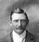 A grainy black and white headshot of a young man with a moustache, dark hair that is both slicked-back and parted, a white shirt, and a dark jacket