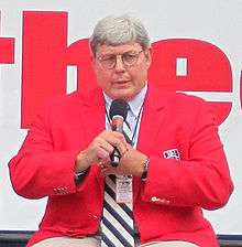 A man with glasses speaks into a microphone wearing a red jacket.