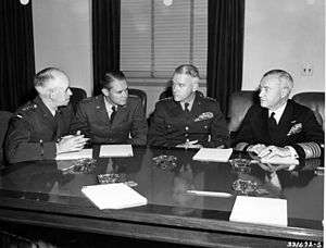 Four men in uniforms but no hats sit at a large polished wood table on which there are pencils, notepads and glass ash trays.