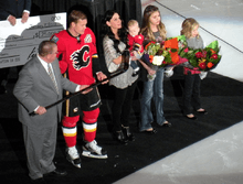 A man in a red hockey uniform accepts a silver stick from another man in a grey suit as his wife and three young daughters stand beside.