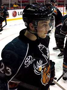 Young male ice hockey player, visible from the waist up, skating while facing towards the right. He is wearing a helmet with a visor, and the uniform logo, a snarling moose, is partially visible on his jersey. In the background three of his teammates, partially obscured, are also warming up.