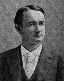 A man with dark hair wearing a black jacket, white shirt, and light tie with a large knot