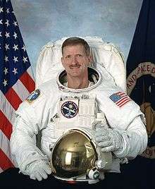 man in space suit holding helmet, American flag in background