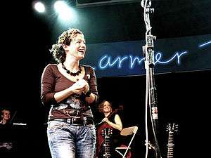 A woman with curly brown hair stands near a microphone stand, wearing a brown T-shirt and jeans.  The necks of two guitars are visible beside the microphone stand.  In the background are two more women, one of whom is holding a cello.