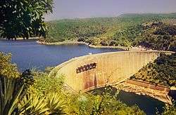 An impressive dam, built in a semi-circular shape, viewed from a vantage point above and to the right of its front. A large lake can be seen on the other side of the dam as well as a river before it.
