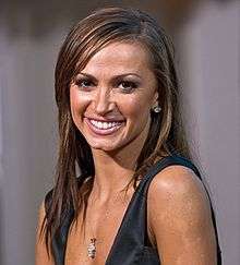 A woman with long brown hair, and tan skin, smiles while looking into the camera.