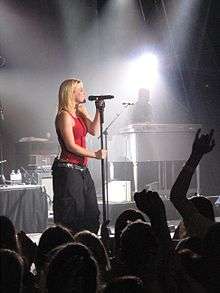 Right profile of a blond woman on a stage. She wears a red top and black trousers and sings in a hand-held microphone. The stage is lit by lights from above and a keyboard player is visible behind the woman. Also visible are the back-profiles of the audiences.