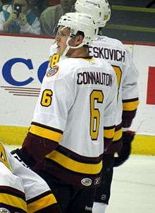 A Caucasian ice hockey player, shown from the waist up, has his back turned to the camera looking over his left shoulder. He wears a white jersey with his name in maroon lettering and the number 6 on his back and sleeve in yellow.