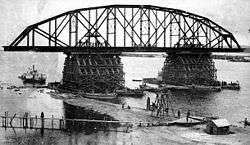 A black and white photograph showing a single truss of a bridge being erected. The steel, semi-circular truss construction is ready and is resting on two pylons erected in the middle of the river and surrounded by scaffolding. Under the bridge a paddle steamer is visible, as well as some wooden temporary causeways and pontoon bridges that allow access to the construction site.