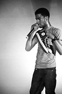 A man clad in a plain T-shirt and jeans speaks into a microphone whilst holding up a pair of sneakers.
