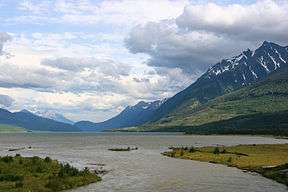 A large body of water, much longer than wide, lies at the base of mountains with vestiges of snow in their higher declivities. Vegetation is sparse. The mountains rise to meet a sky filled mostly with puffy white or gray clouds.