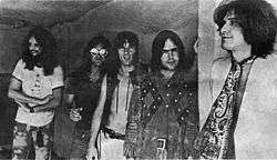 Five smiling men in a row, diagonal to camera angle. The man on the left (farthest to the back) has very long hair and a full beard; he wears a white T-shirt and tie-dyed pants. Next to him, Dave Davies, also with very long hair, wears reflective sunglasses, a black short-sleeved shirt, and jeans. In the middle, Mick Avory wears an unbuttoned leather vest and white pants. The man to his right wears a heavy, probably brown leather jacket with a design that is possibly Native American. On the far right, in front, Ray Davies wears a giant paisley kerchief knotted like a tie, over a white jacket.