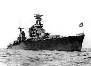 Water-level bow view of a large grey ship flying the Soviet Naval Ensign. Two gun turrets and the forward superstructure are prominent.