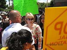 A woman in her thirties with fairly short blond hair, wearing sunglasses and a beige and pink top, is surrounded by a crowd in an outdoor setting. Two signs are being held by the crowd, one with Bible quotations about loving others and the other depicting a stop sign. On the right is the corner of a building labeled State Senate.