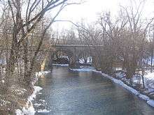 A winter view of a stream with snow on the banks. A stone and concrete railroad bridge appears near the center of the photo, crossing the stream. The trees are bare and power lines appear near the center of the photo as well.