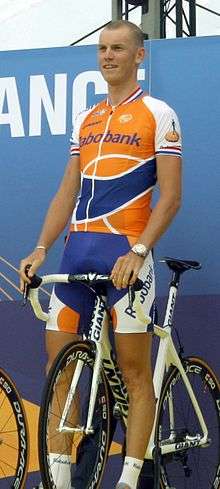 A road racing cyclist in an orange and blue jersey with white trim, standing over his bicycle which is stationary.