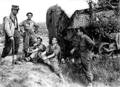 A man in uniform carrying a walking stick and wearing a kepi talks to four men in overalls wearing berets. A heavily camouflaged tank is in the background.