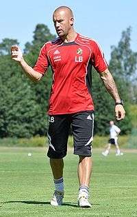 A photograph of a bald man on a training pitch wearing a red sports sweater, black shorts and white socks.