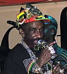 A man singing into a microphone; he is wearing a colorful hat with many accessories on his hat and around his wrists, fingers and neck.