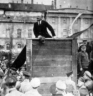 A black-and-white photograph of a crowd scene.  A bald, goateed man stands on a platform in the centre-left, speaking dramatically to the crowd.