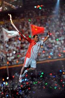 An Asian man in red and white athletic shirt and shorts, and wearing athletic shoes, is suspended by wires in the air while holding a lit torch. In the background, a large crowd in a stadium can be seen, as well as two blurred flags.
