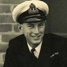 Monochrome photograph of head and shoulders of smiling young man (Lieutenant John Paul Wild RNVR) in white Royal Navy officer's cap and naval uniform.